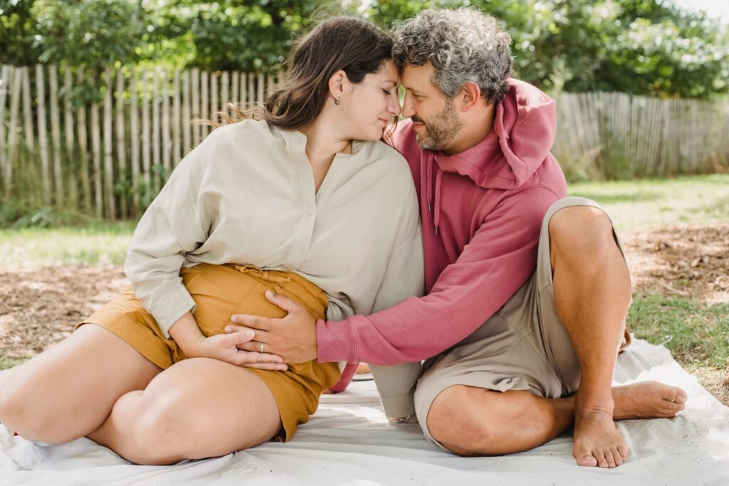 Side view of tender husband touching belly of pregnant wife while sitting on blanket and touching with foreheads in countryside
