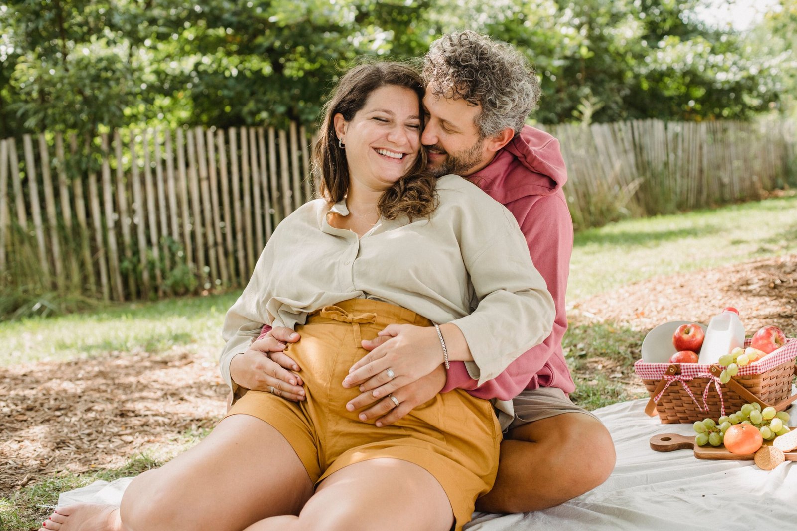 Loving pregnant woman and husband with closed eyes sitting on blanket with hands on belly while hugging during romantic date in countryside